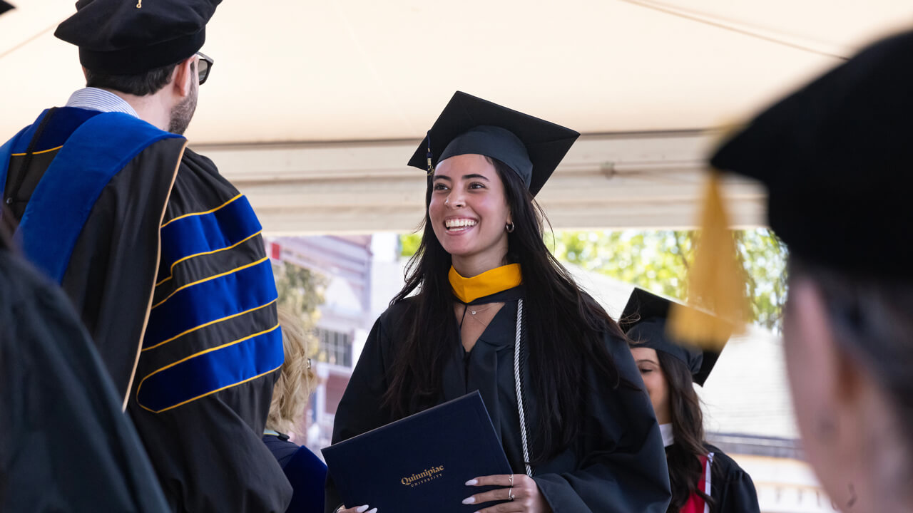 A graduate holds her diploma and smiles on the library steps