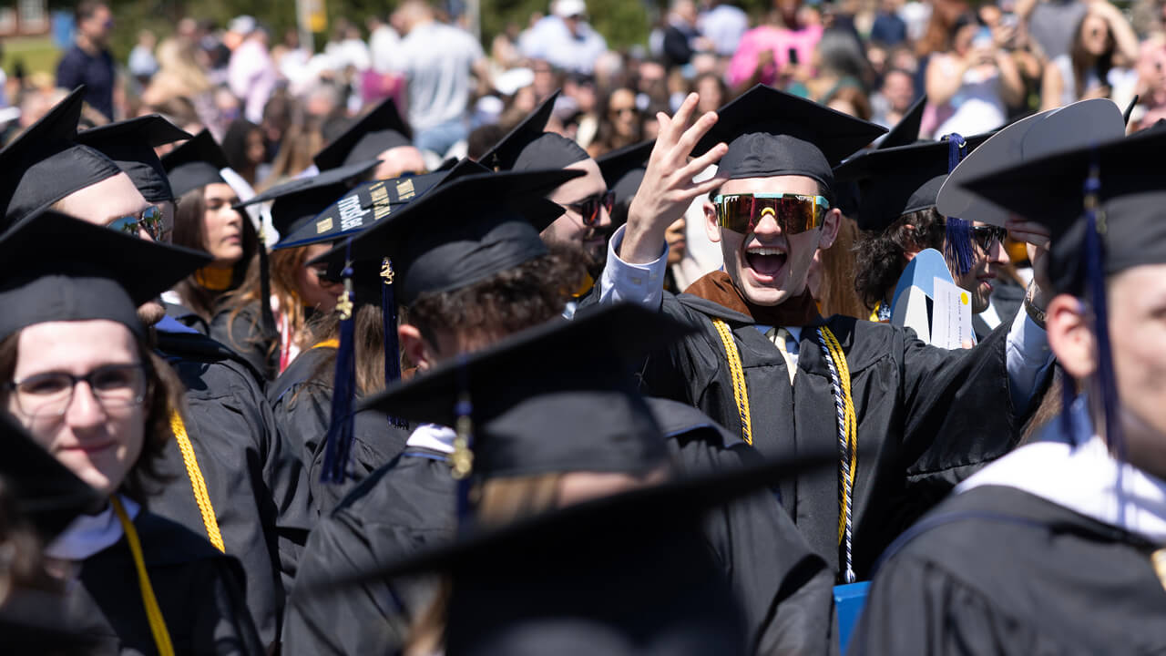 A graduate cheers while wearing shiny, reflective sunglasses as he sits