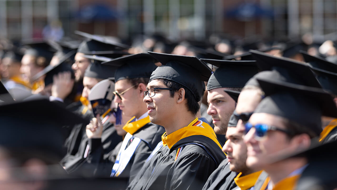 Graduate wearing glasses looks towards the stage while seated in the crowd