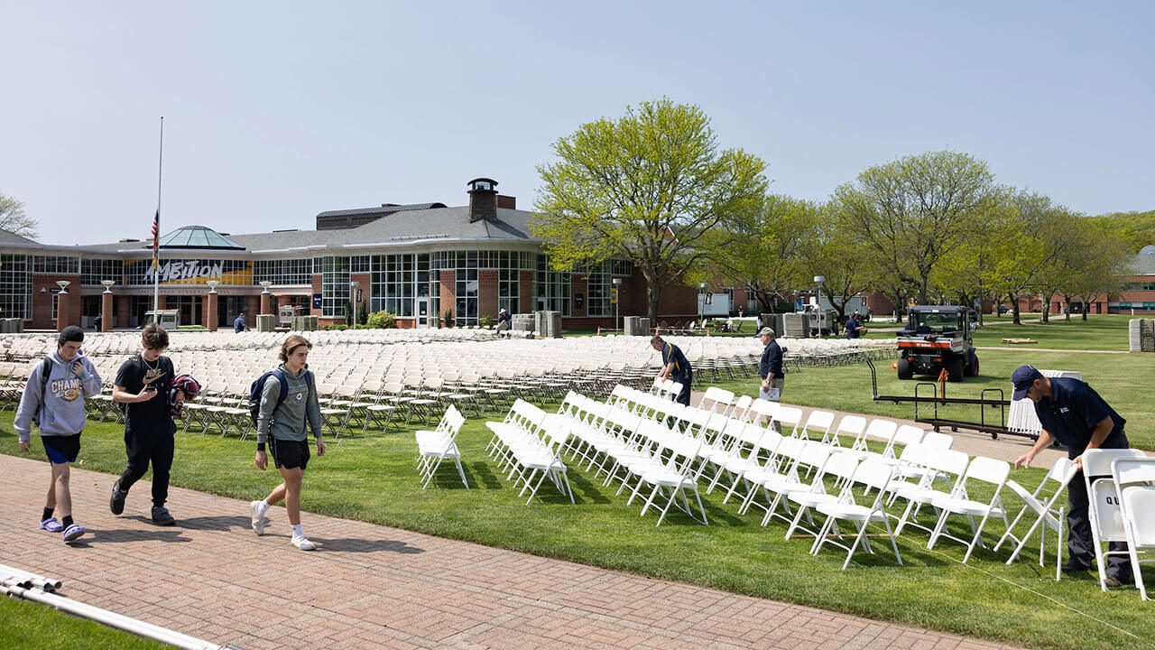 facilities employees unload white chairs while three male students walk past