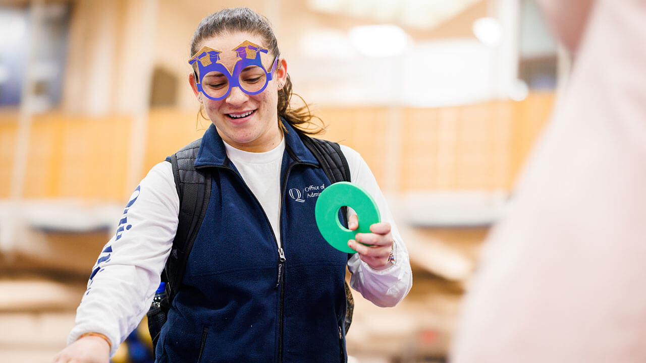 female student with a ponytail and purple glasses with graduation caps on them holds a green O in her hand as she reaches for something on the table