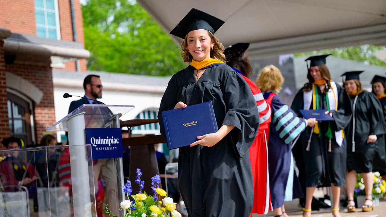 female graduate walks away from receiving her diploma with a huge grin on her face as she holds up her diploma to the camera