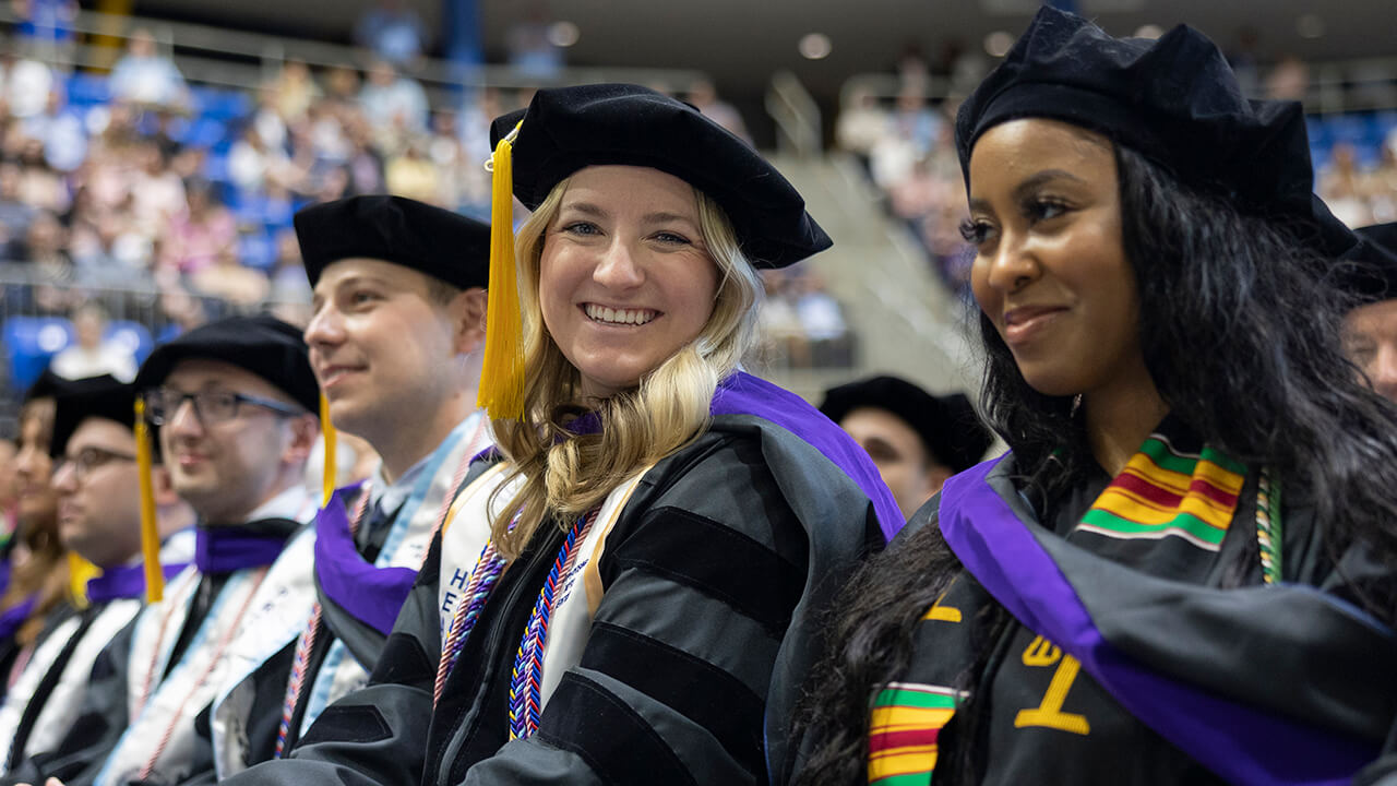 Graduate smiles at the camera while seated with colleagues