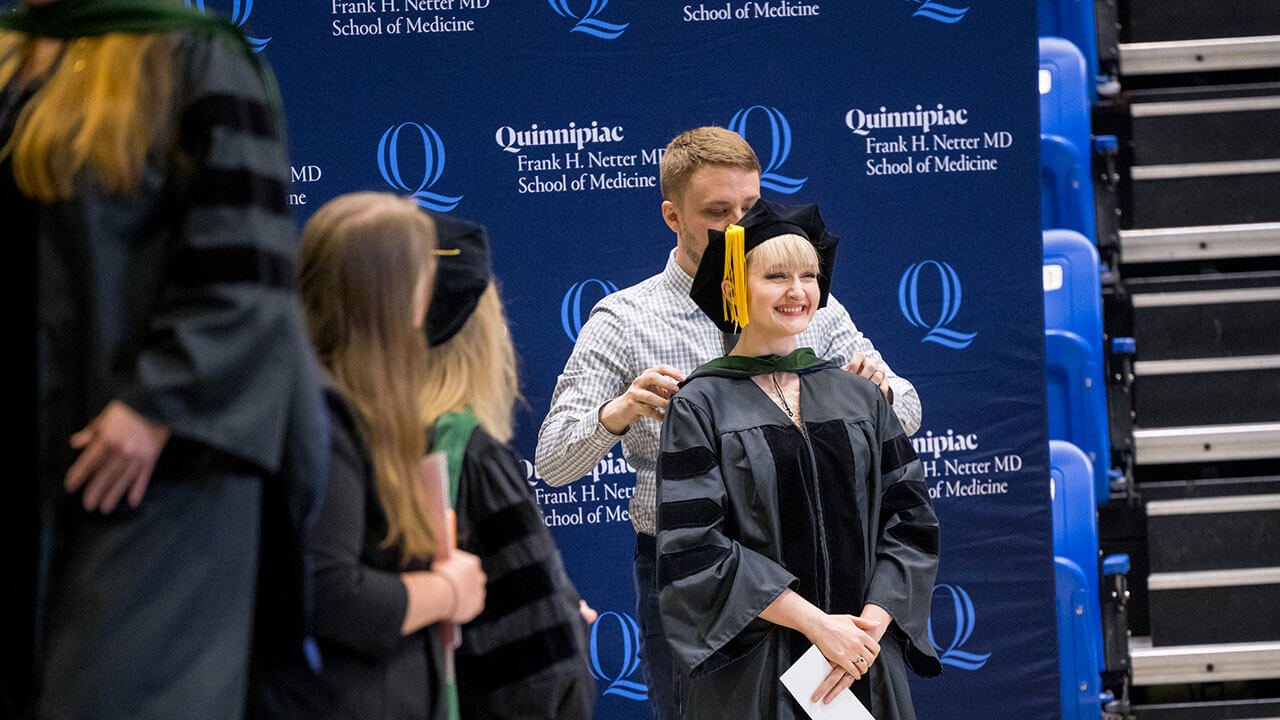 a blonde female medicine graduate is hooded by a man in a blue button down shirt and tie