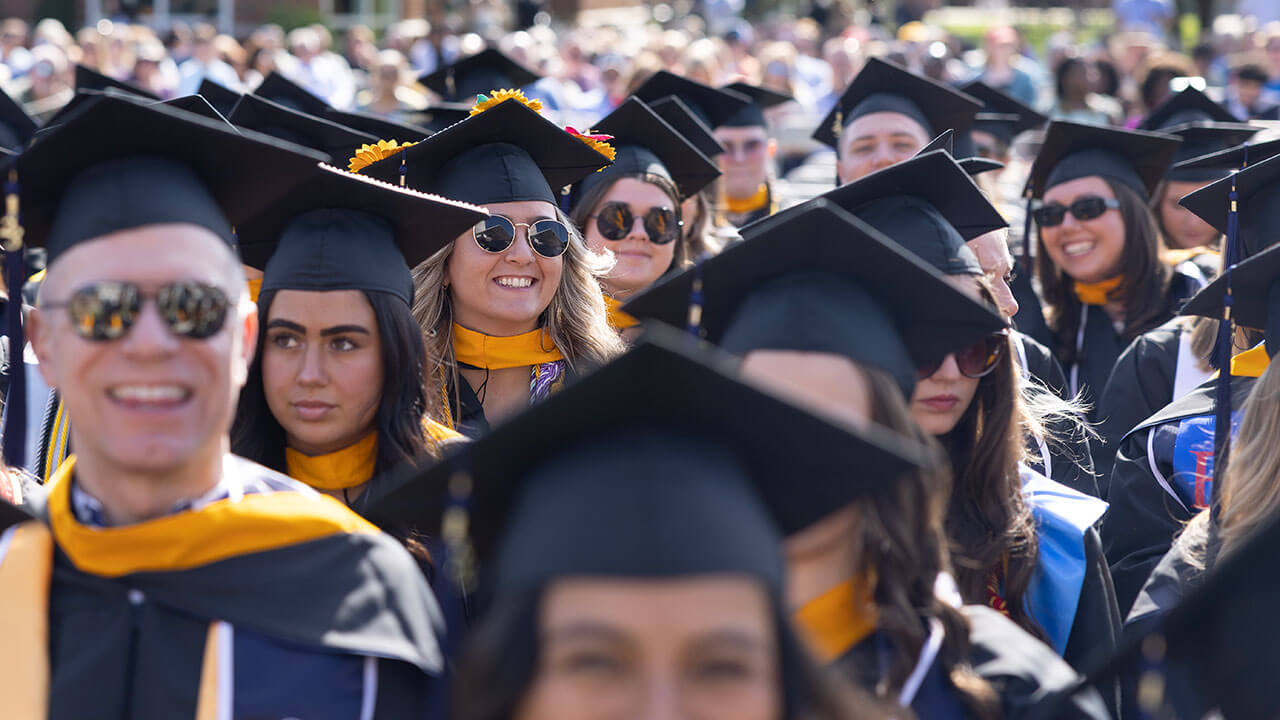 A sea of graduates in sunglasses sit and smile broadly in the sunshine