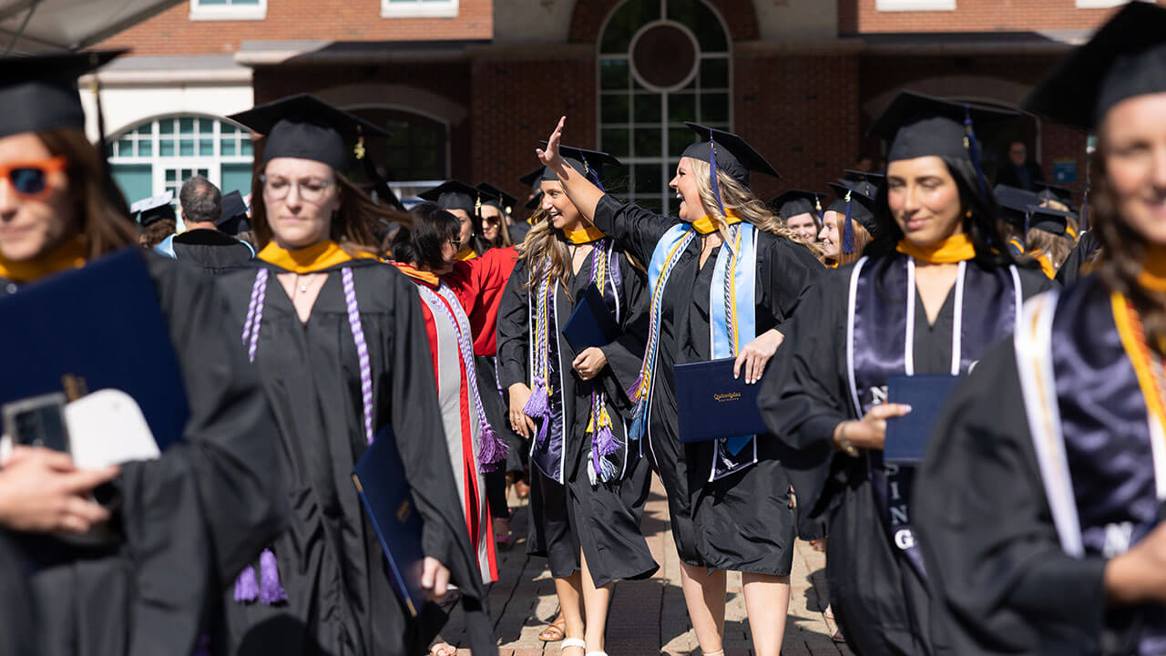 graduate walks down the center aisle and smiles broadly waving at an audience member