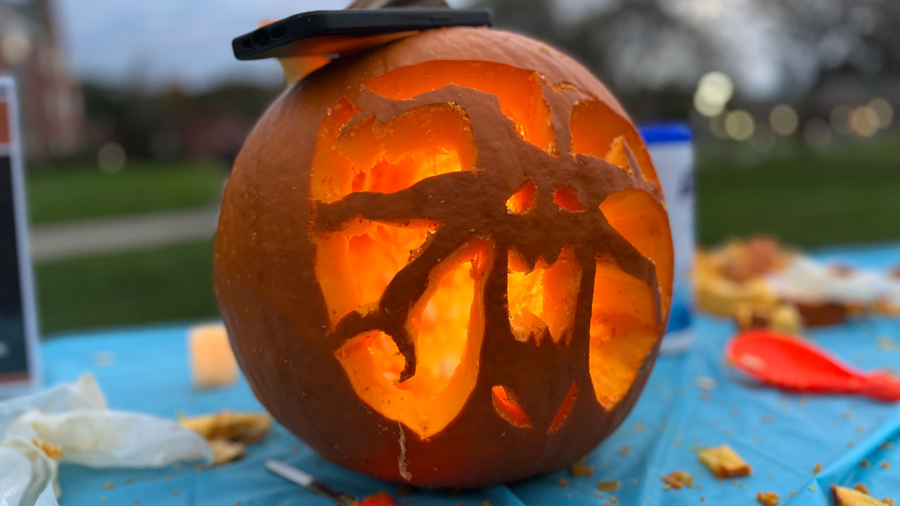 A pumpkin carved into the shape of a tree with a face sits on the table with a phone flashlight shining through to illuminate it