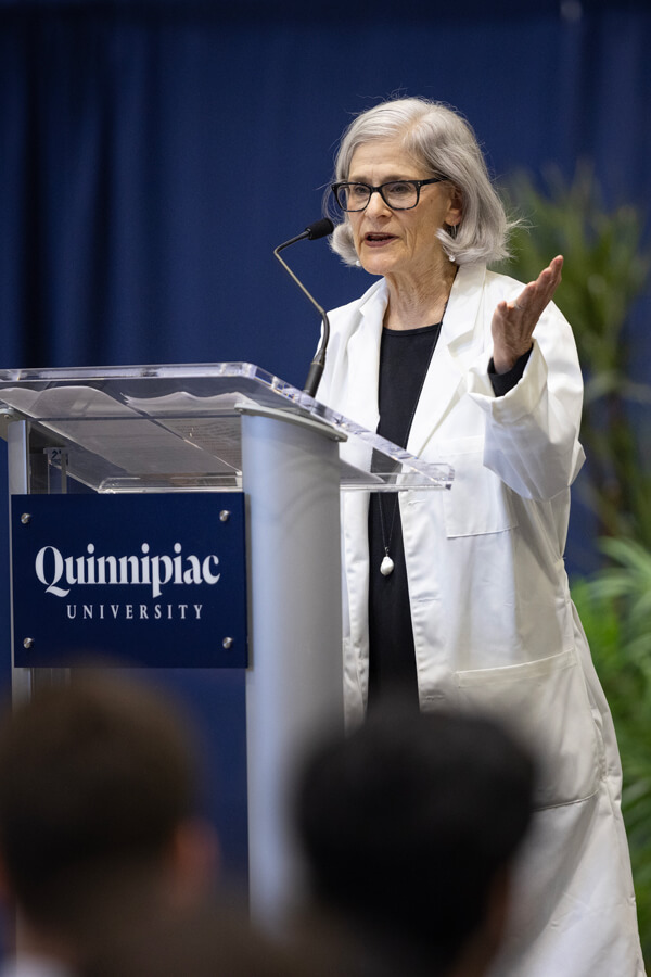 Katherine Trymbulak wears her white coat and speaks from the podium with green plants behind her