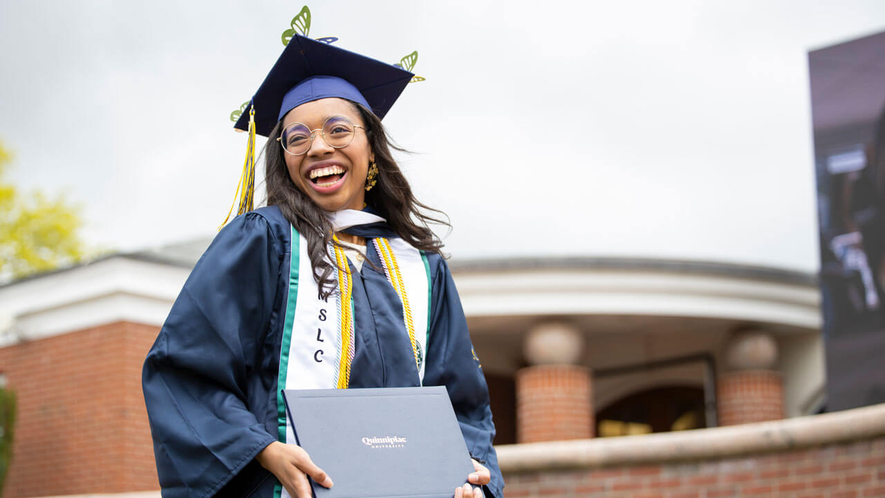 A beaming College of Arts and Sciences graduate proudly holds up their diploma while wearing a graduation cap decorated with butterflies.