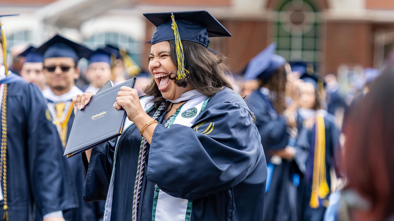 A graduate enthusiastically smiles and holds up her diploma in victory