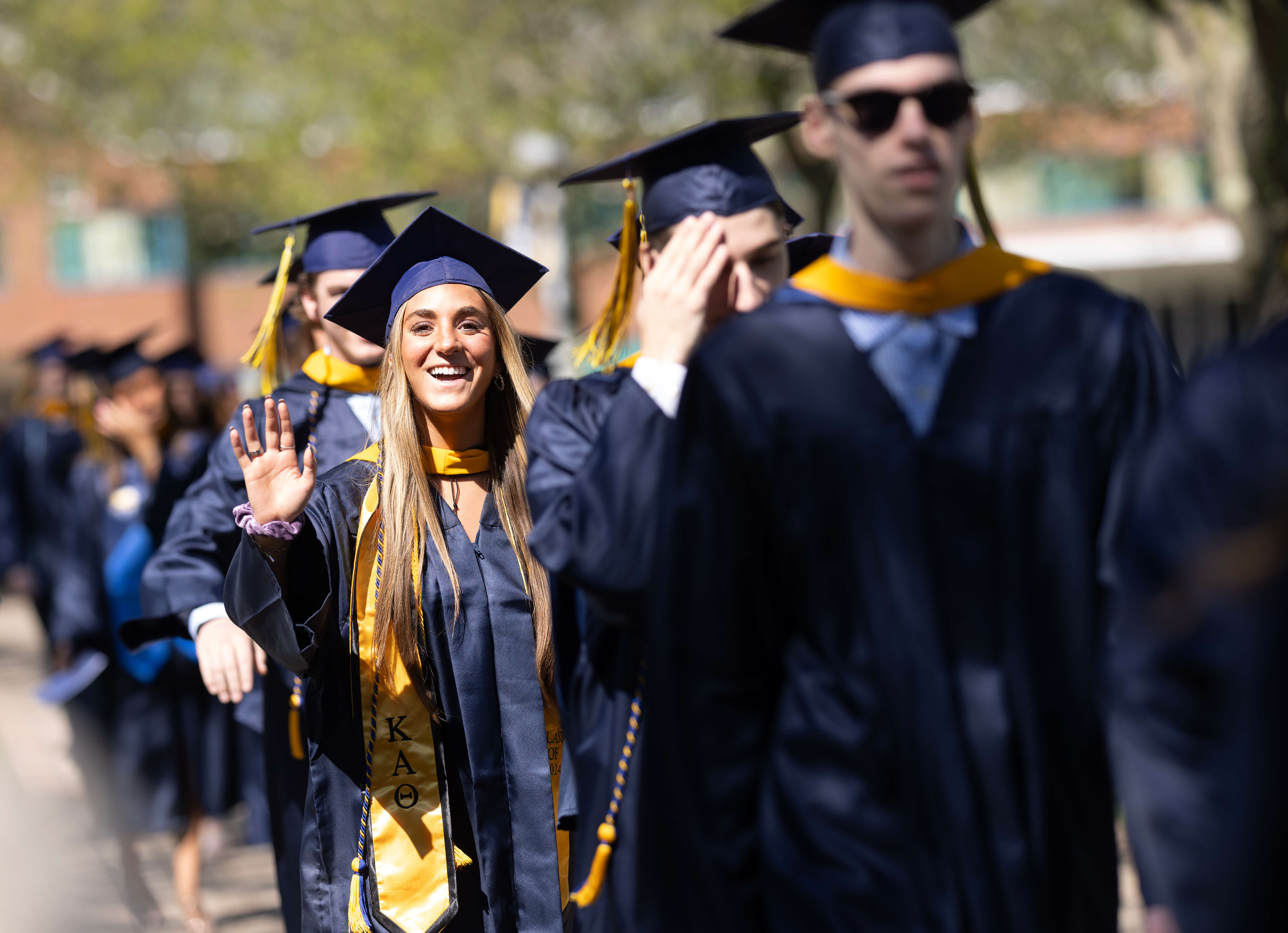 Graaduates walking in a line to their seats, one girl waving to the camera