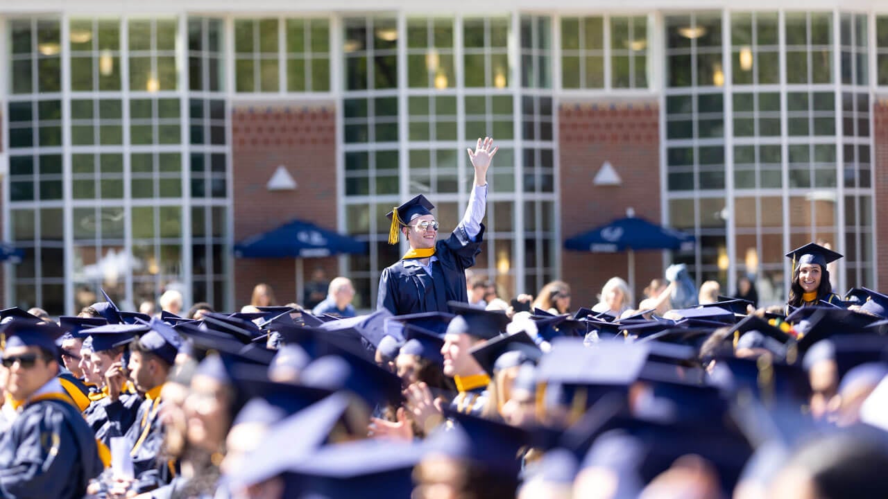 Student in gap and gown stands up in crowd to waive