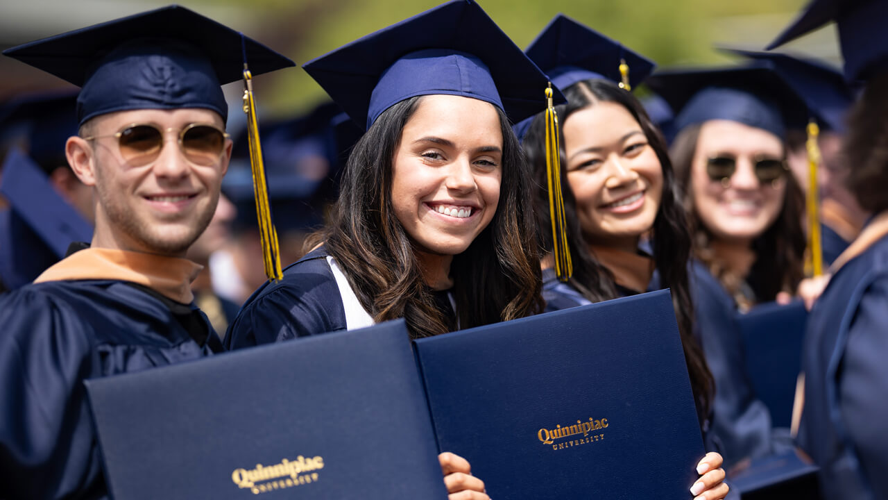 Four students sit in a row in their seats and hold up their diplomas and smile