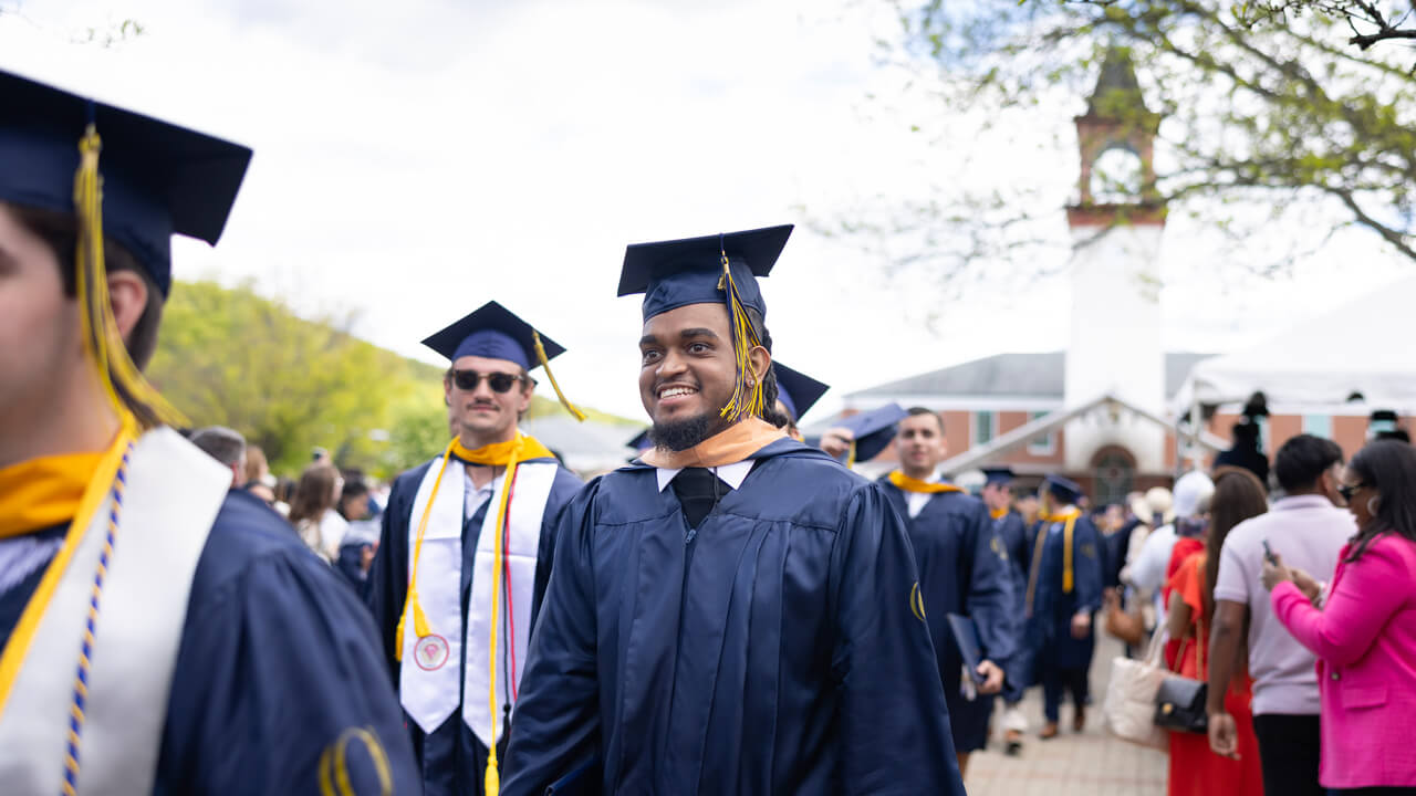 Dozens of graduates walk past guests on the quad with the clocktower behind them