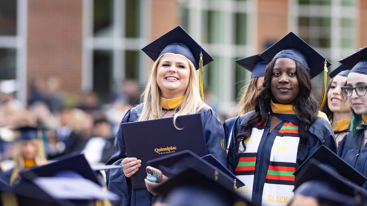 Five graduates stand and hold their diplomas behind dozens of seated graduates