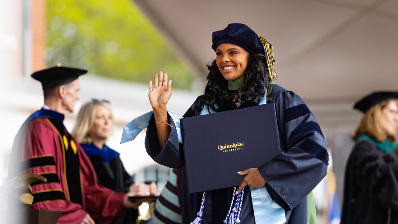 A graduate smiles and waves as she holds her diploma and crosses the stage