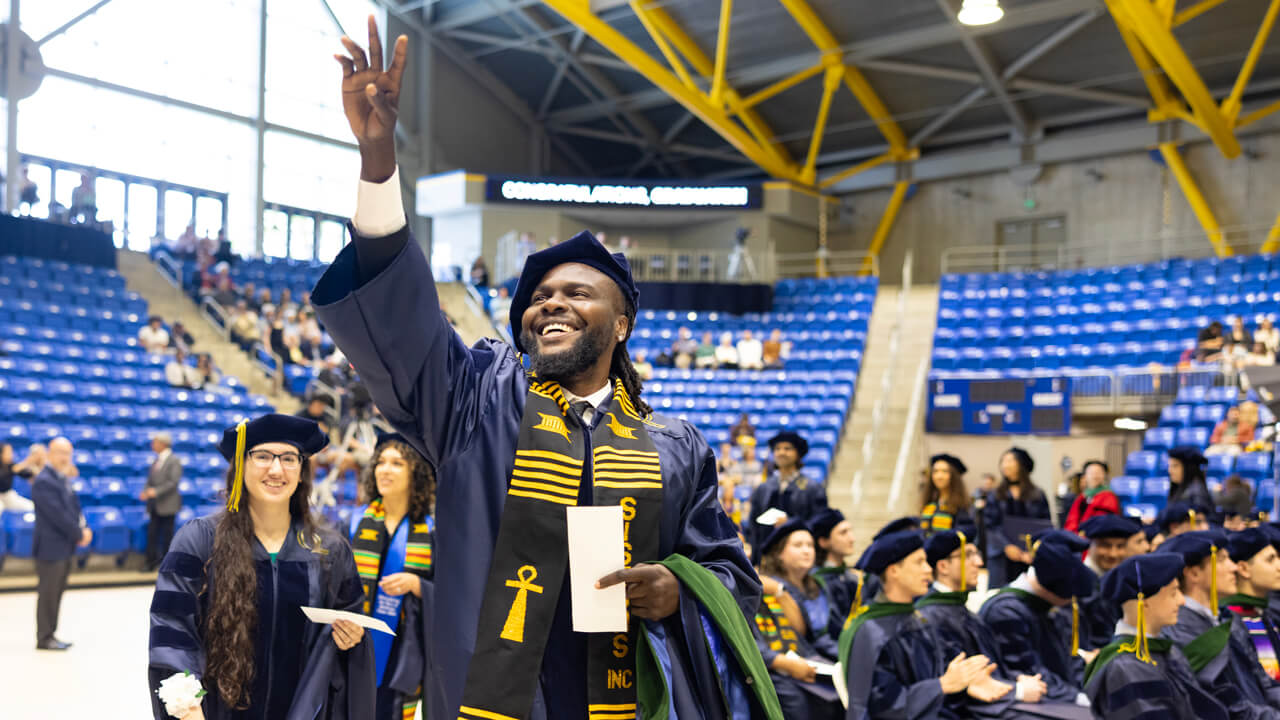 A graduate smiles broadly and waves to the audience as he walks into the ceremony