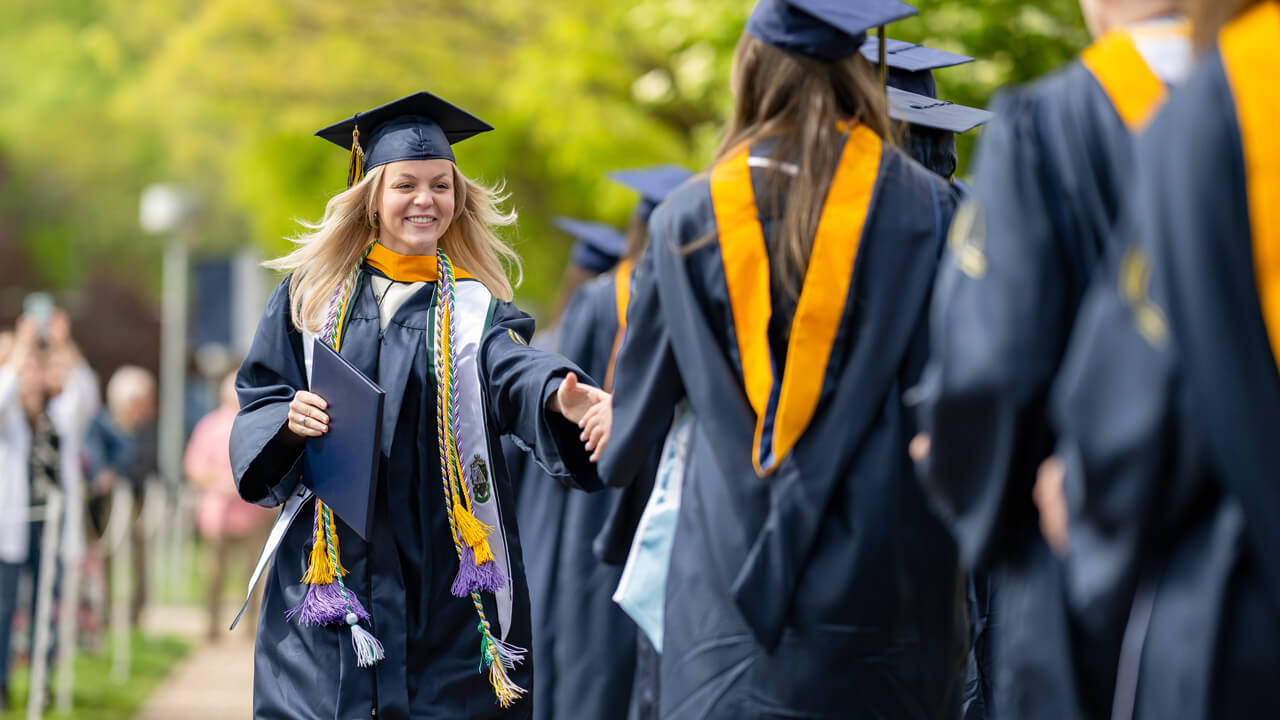 A graduate wearing a white stole smiles and reaches out her hand