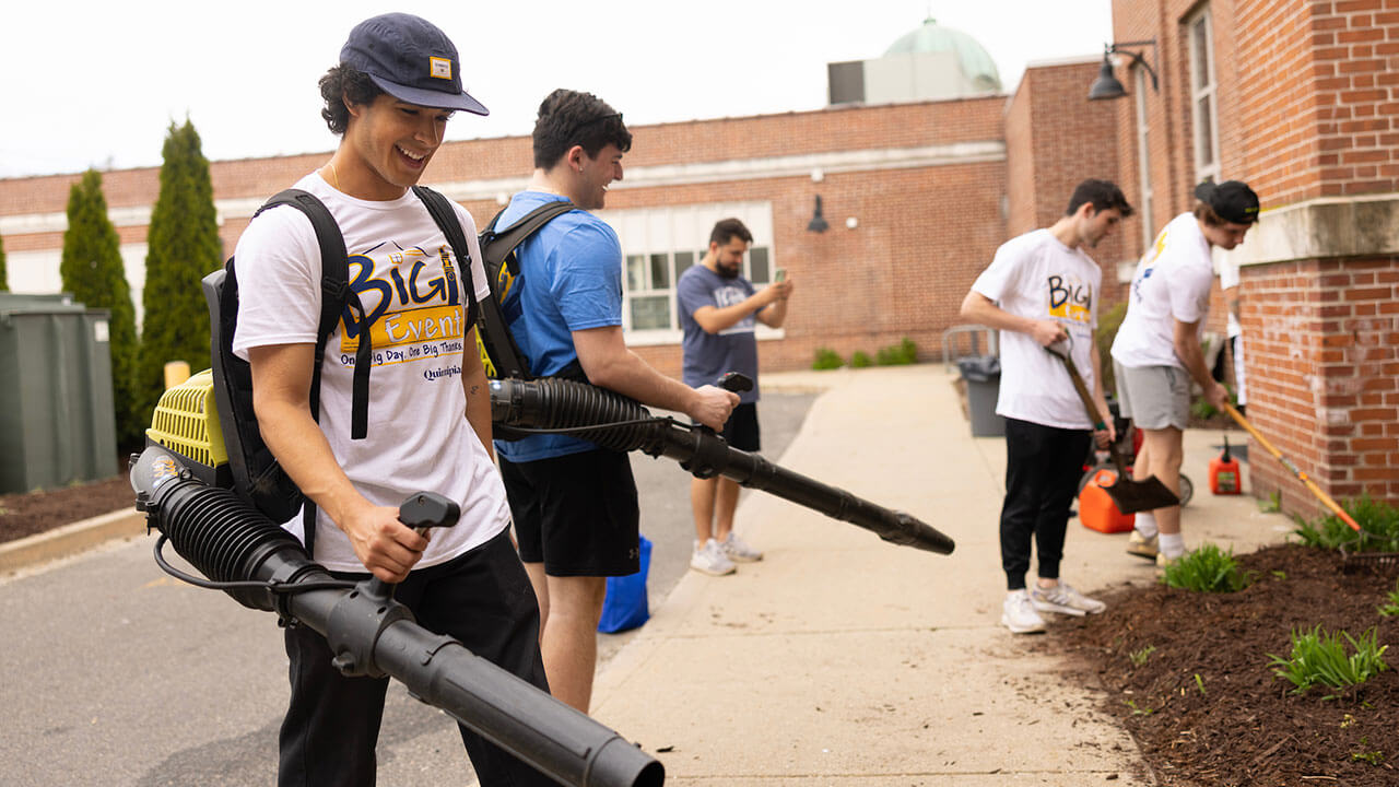 two male students in focus use leaf blowers to clean up loose mulch from the sidewalk, two out of focus students move mulch into place