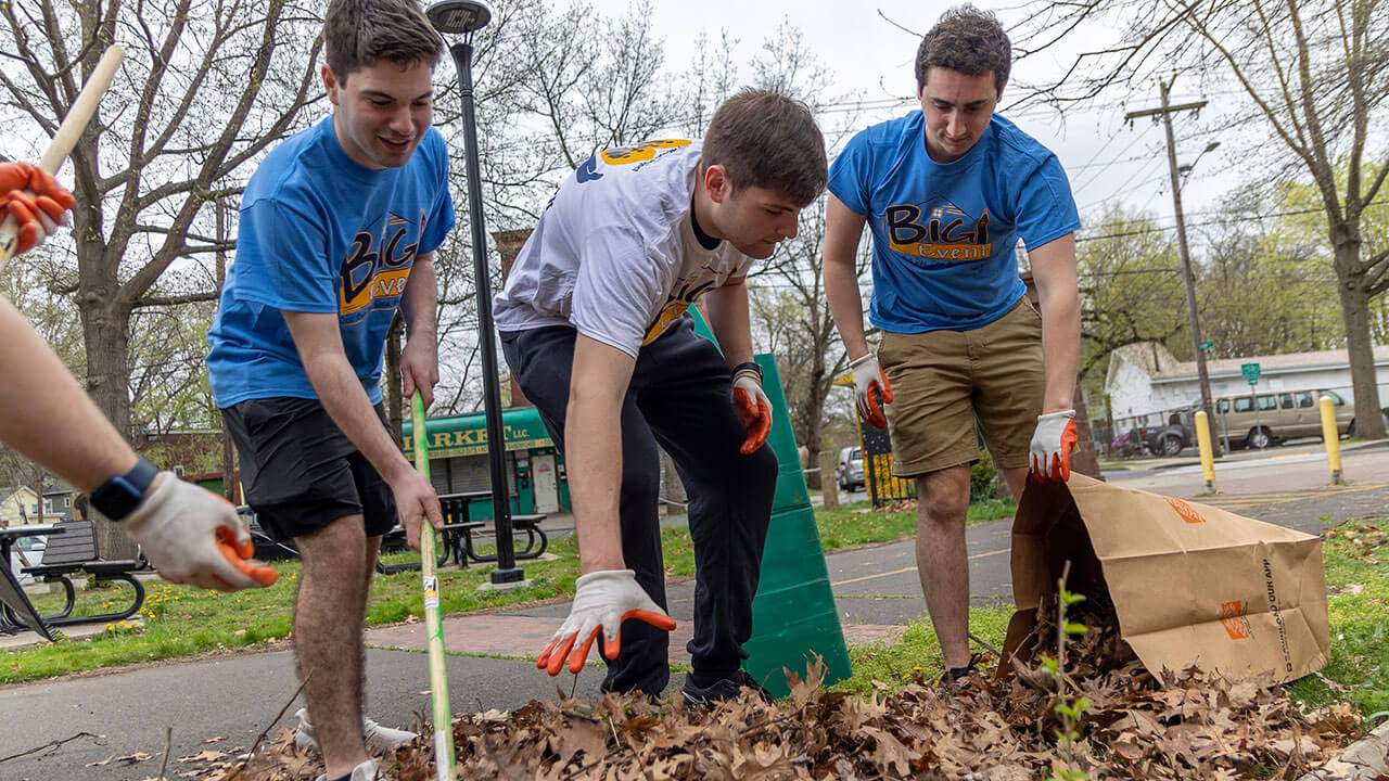 three smiling male students rake leaves into a paper bag, grabbing fist fulls of the leaves