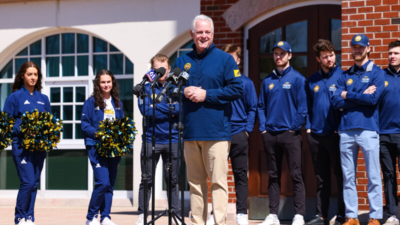 Athletic director Greg Amodio speaks at microphones in front of the men's ice hockey team