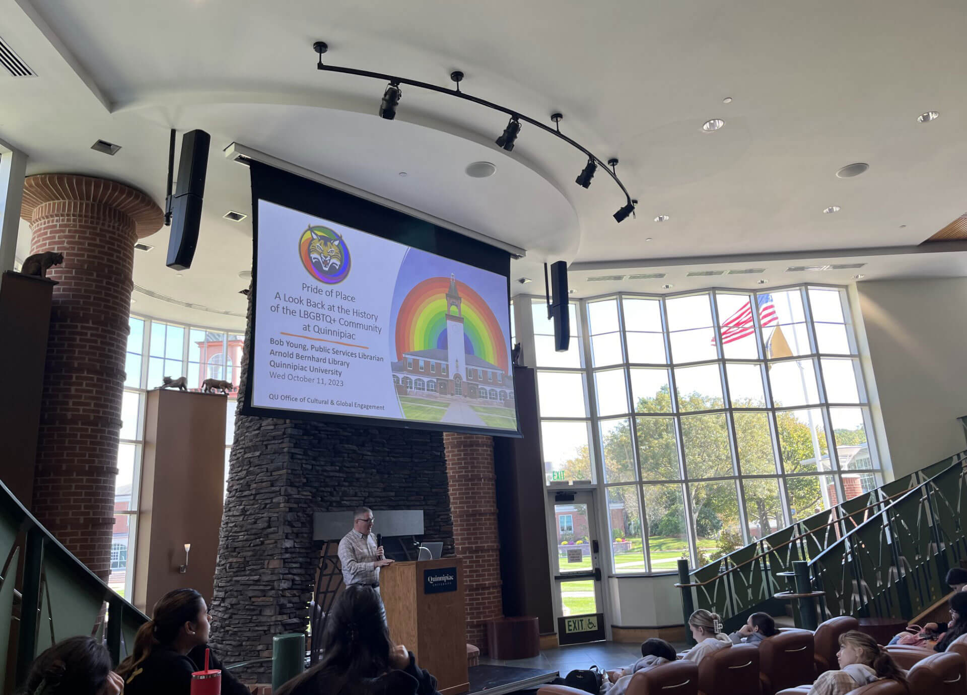 People sit in the Piazza to watch presentations at the LGBTQ teach-in where a speaker stands on the stage.