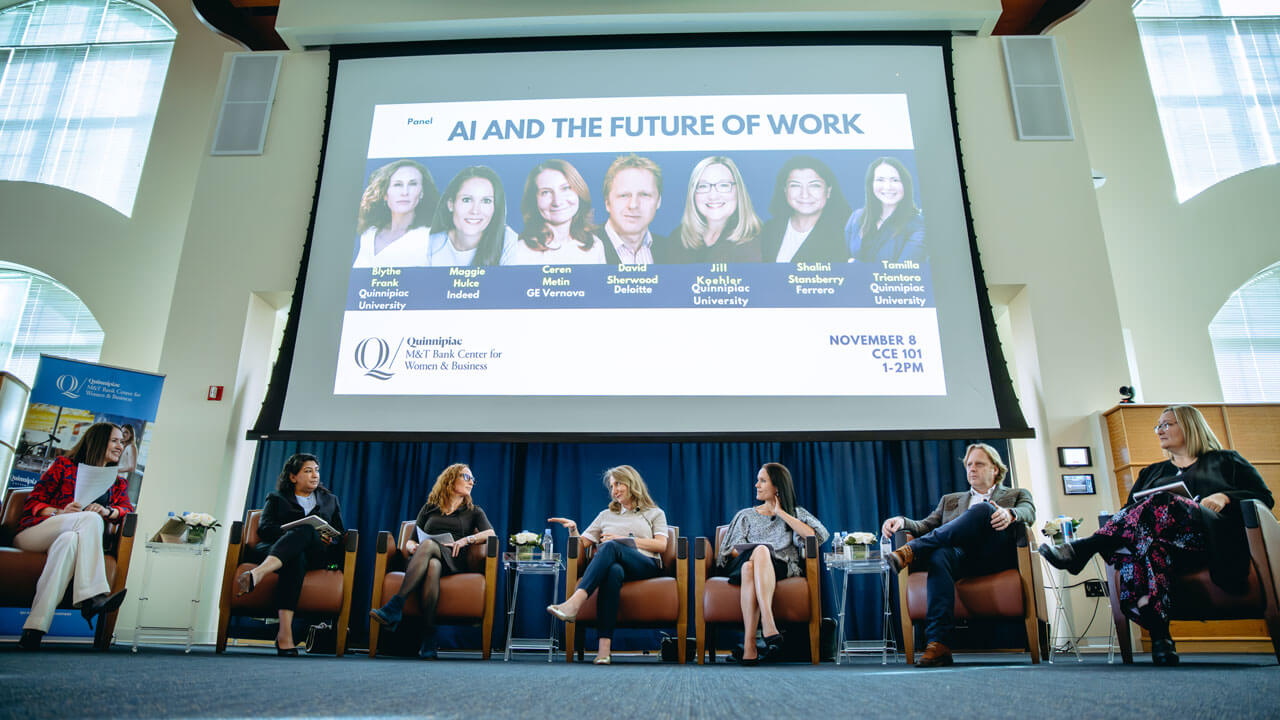 A group of panelists sit in chairs talking with a large screen behind them.