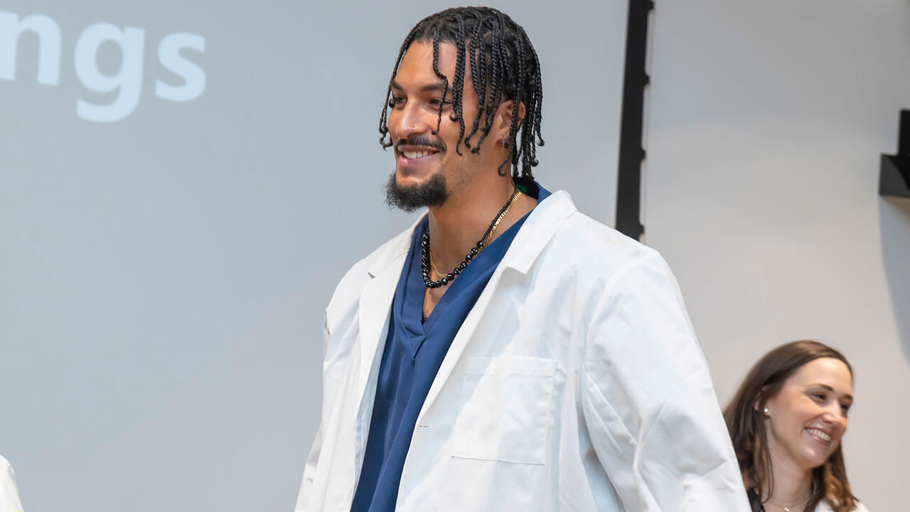 nursing student has his white coat on as he walks towards the front of the room