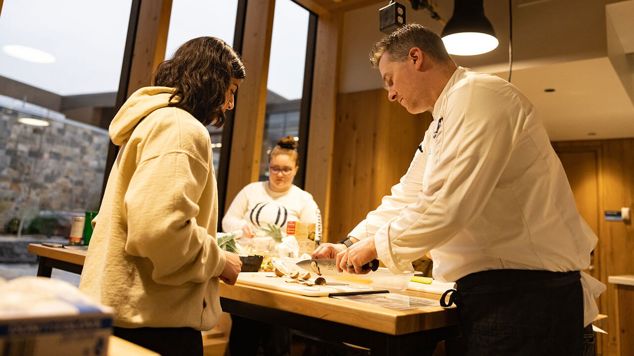 chef demonstrates how to cut up a mushroom for a student with short dark wavy hair