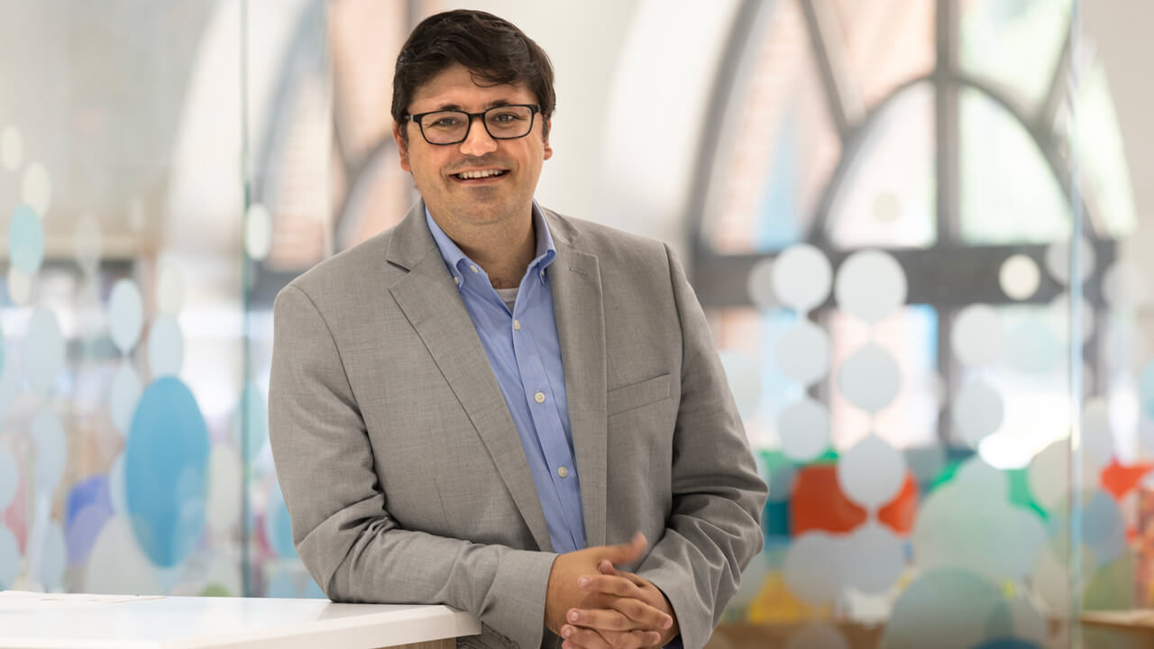 James Demetriades leans on a small wall for a photo in front of a wall of colored glass