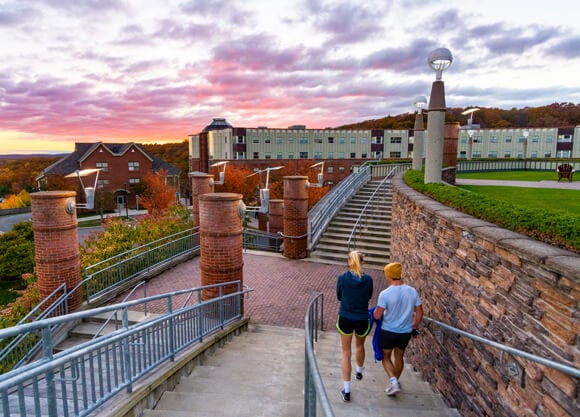 Two students walk down stairs below a pink and gold sunset and Long Island Sound in the distance