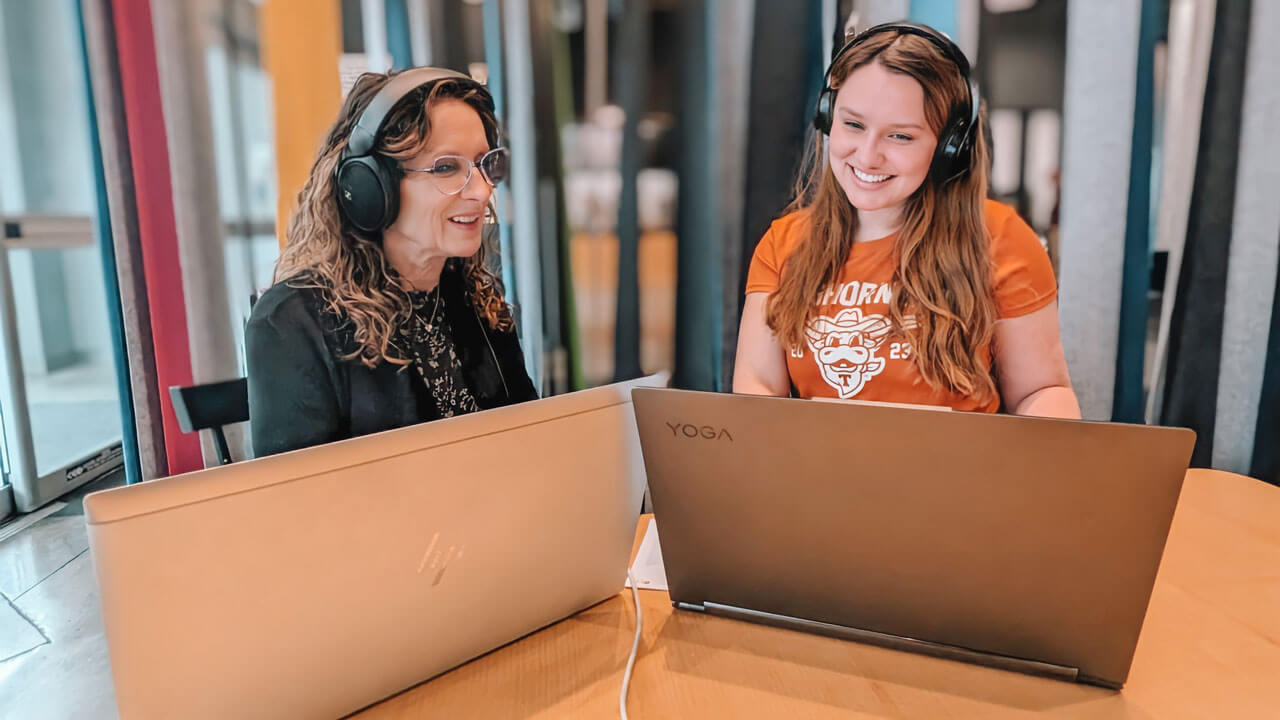 Kathryn and Tesni sit behind their computers smiling and wearing headphones.