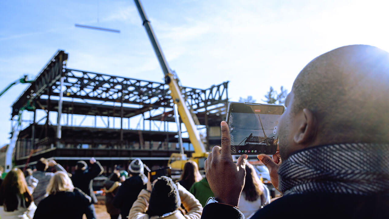 An individual takes photos of a beam being placed on a building on the South Quad.