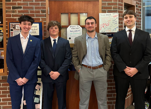 Four students dressed in suits stand together and pose.