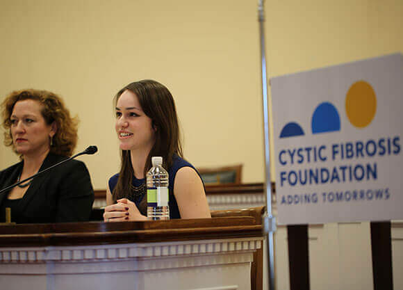 Cray sits at a table and speaks in to a microphone.