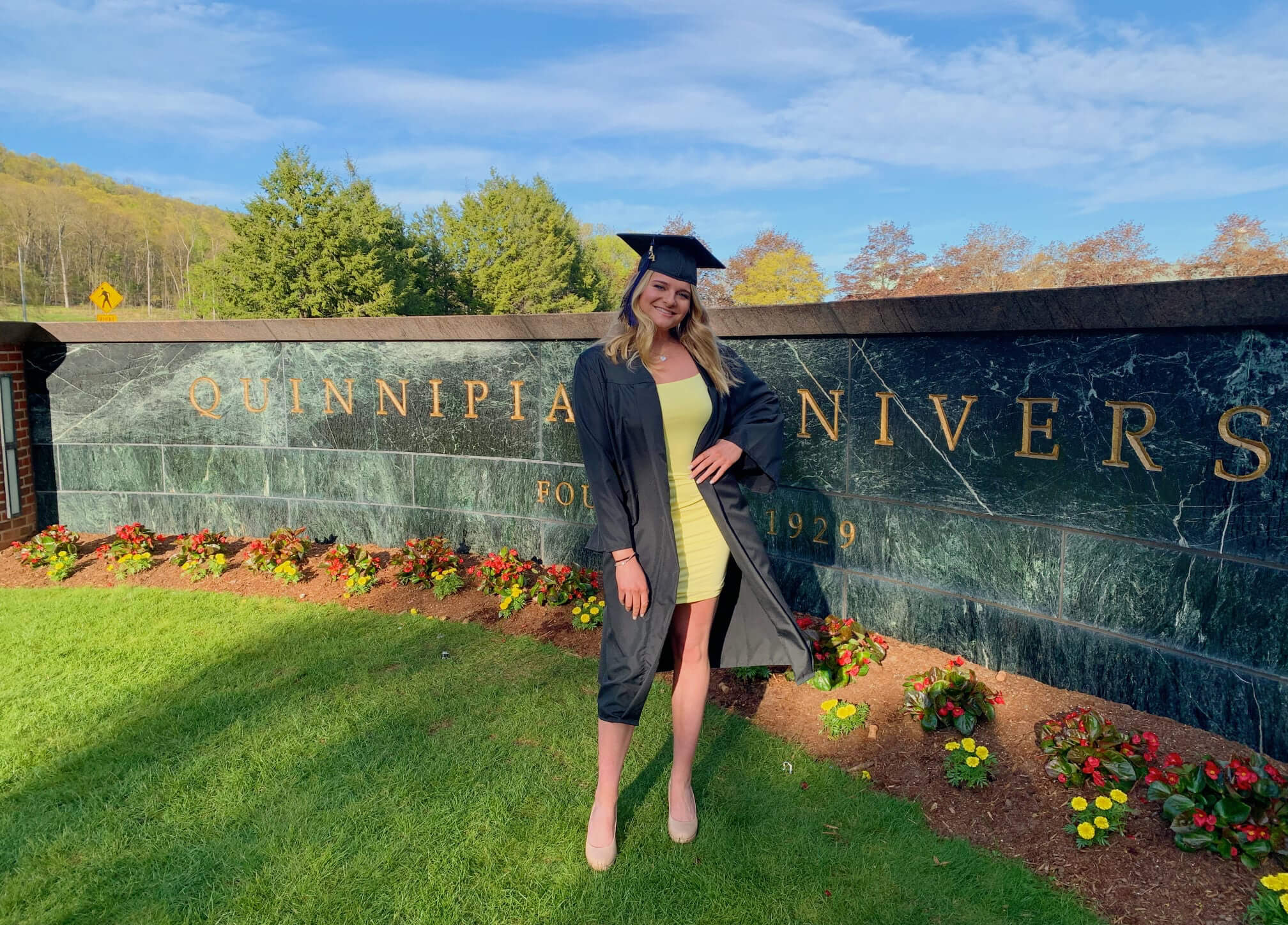 Heather Popovics standing in front of Quinnipiac sign at Mt. Carmel campus in graduation regalia