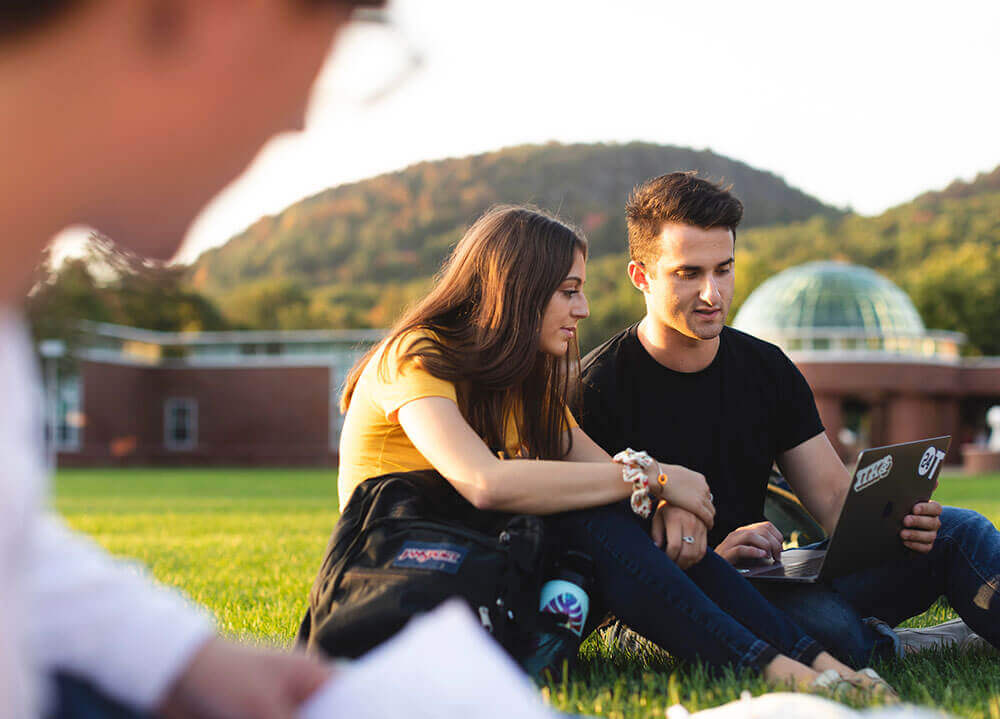 Two students sit on the Mount Carmel Campus quad sharing a laptop with a view of Sleeping Giant park in the background