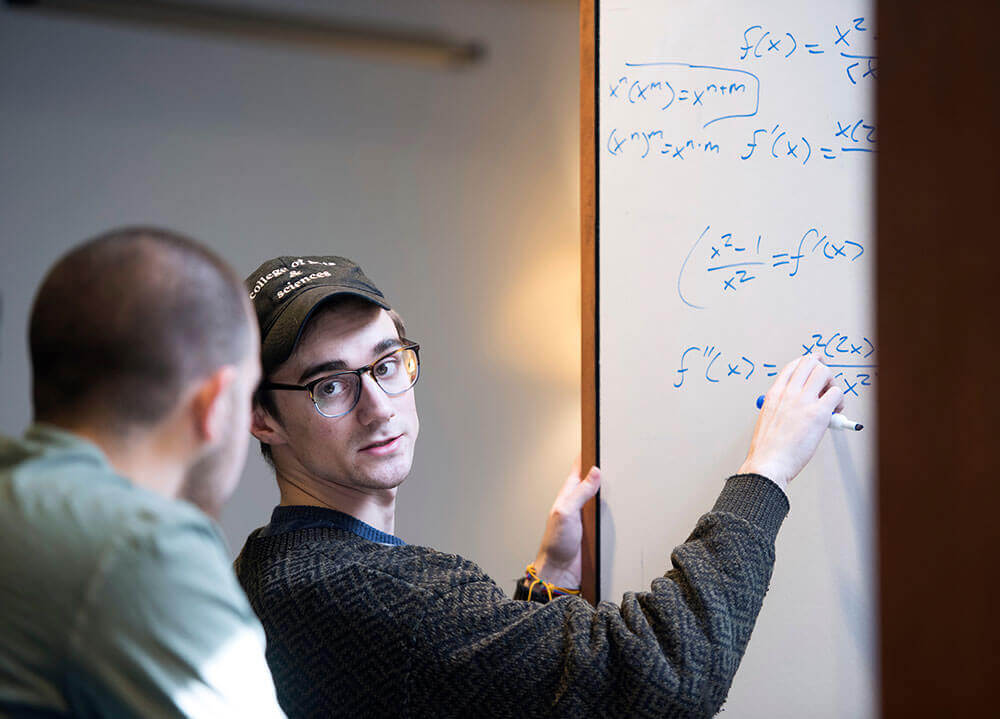 A student tutors another student in math by writing equations on a white board with blue marker