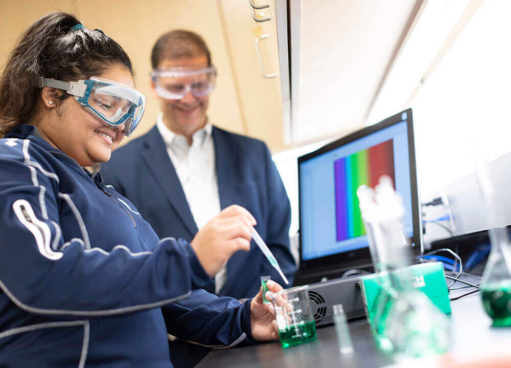 A biochemistry student conducts a laboratory procedure while her professor looks on in the background