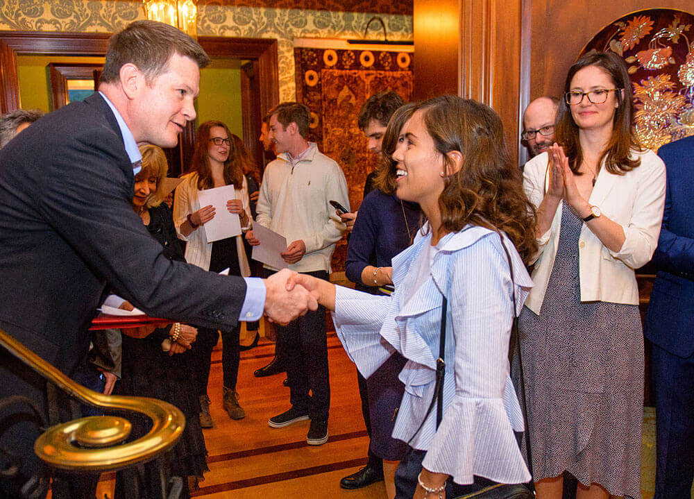 An economics student shakes a professor's hand during a formal networking event at a trustee's home