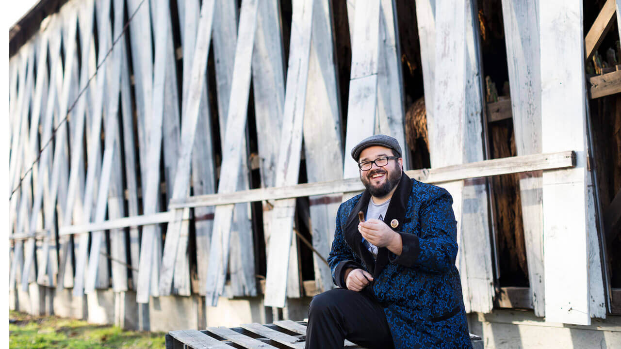 Nicholas Melillo smiles holding a cigar while sitting in front of one of his tobacco barns