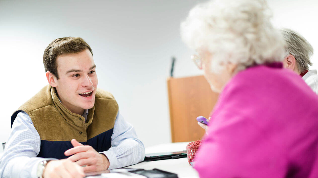 A student speaks with an elderly woman while they sit across from each other at a table