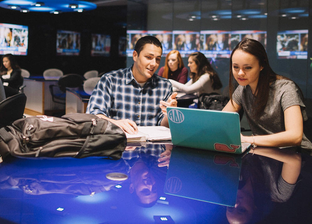 Two communications students collaborate on coursework sitting next to one another with their laptops