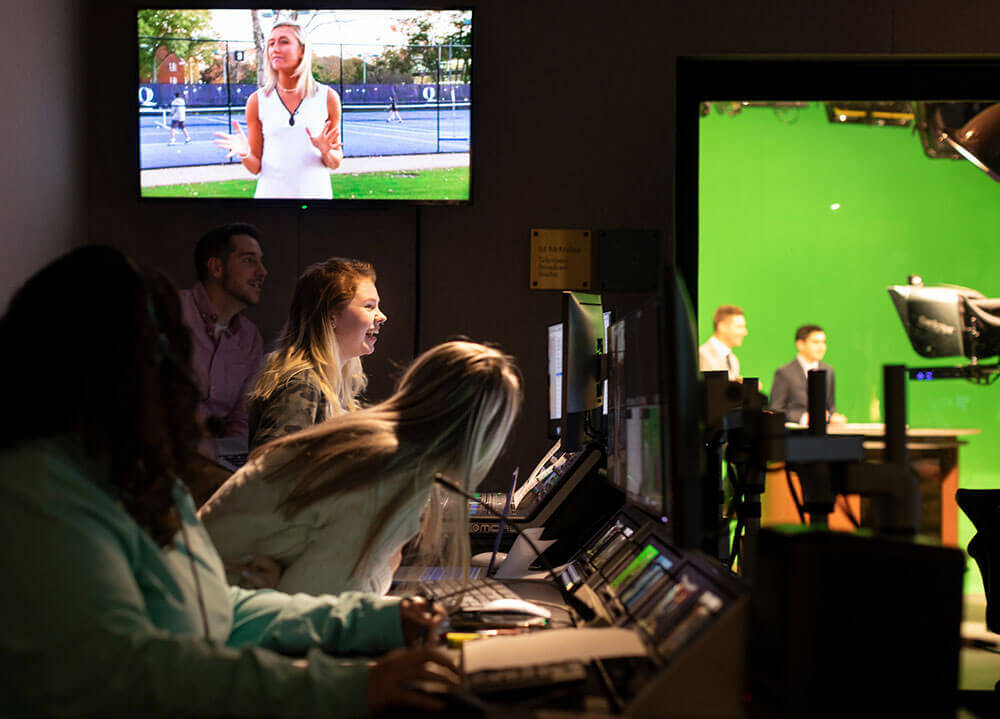 A group of students and a professor sit behind large master control boards in the control center of a live news broadcast