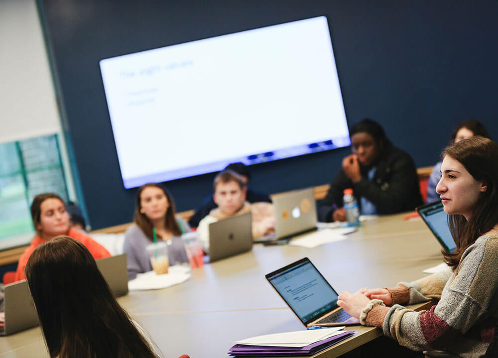 A group of students sit around a classroom table with their laptops open