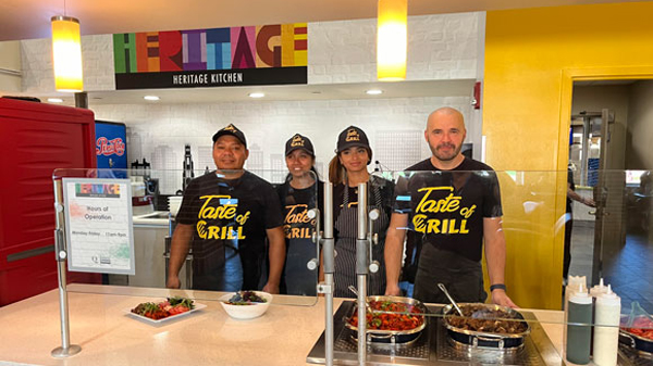 Three cooks stand behind a counter with their cuisine and a sign that reads Heritage Kitchen