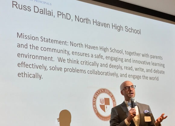 North Haven High School principal Russ Dallai gestures while holding a microphone in front of a screen.