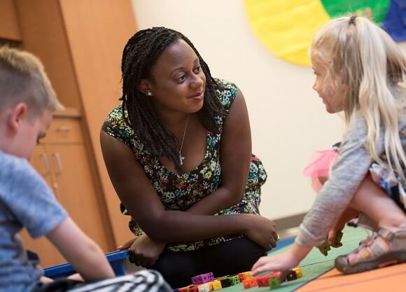 Student kneels down to interact with two children on a colorful mat