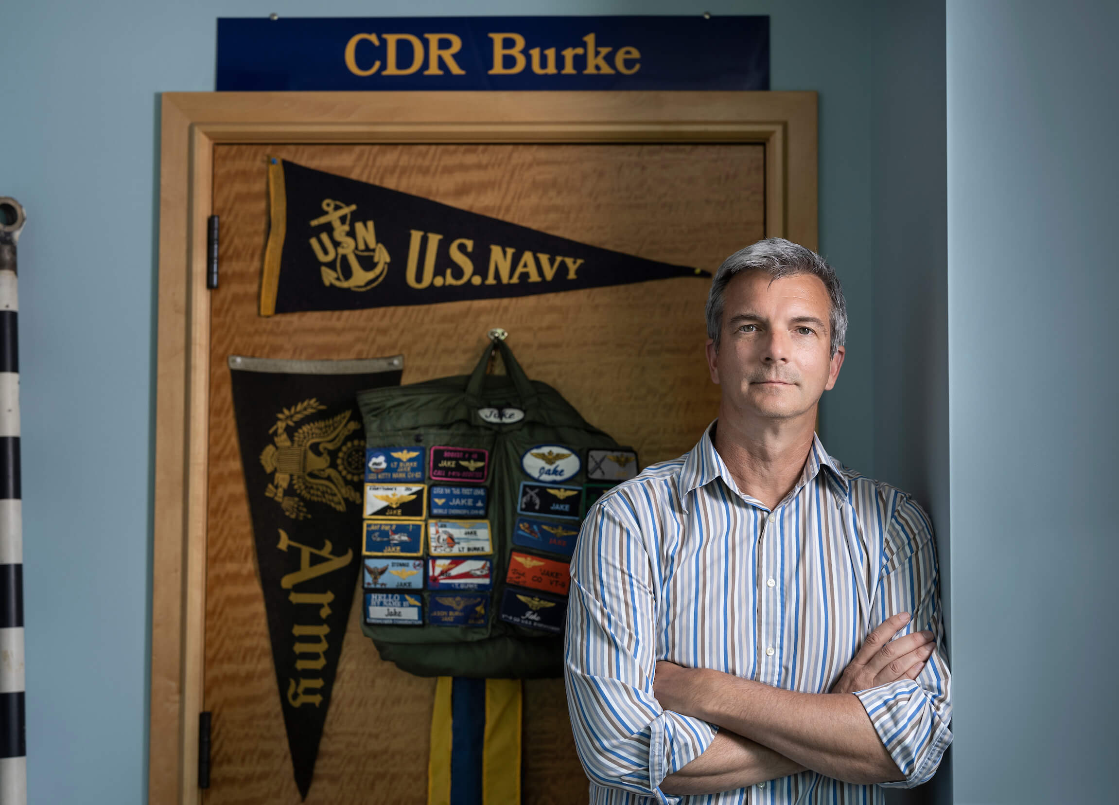 Jason Burke in his home standing in front of US Navy flag and backpack with patches